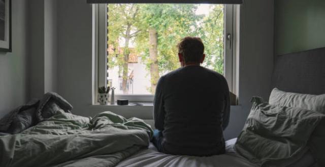 homme assis sur son lit regarde à travers sa fenêtre